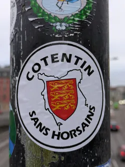 Street sticker A round sticker with the text "COTENTIN" at the top and "SANS HORSAINS" at the bottom.  In between is an outline of the Cotentin Peninsula with a coat of arms featuring golden lions on a red shield. The sticker is affixed to a dark-colored pole.
