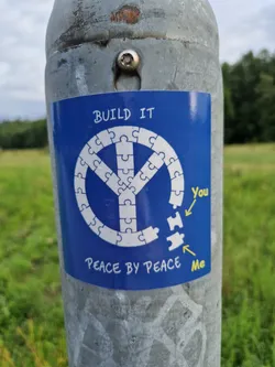 Street sticker A blue sticker with a peace symbol made of puzzle pieces. The text 'BUILD IT' is at the top, and 'PEACE BY PEACE' at the bottom. There are two missing puzzle pieces labeled 'You' and 'Me'.