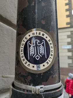 Round sticker with a dark blue center and beige outer ring. The central design features a stylized eagle with wheat stalks, and text in a circular pattern reads "Vereinigte Bier Republik" which translates to United Beer Republic. A QR code is visible at the bottom.