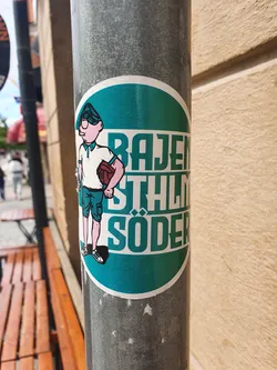 Street sticker A teal round sticker with a cartoon illustration of a person holding a ball and text that says "Bajen Stålhlm Söder". The sticker is affixed to a gray metal pole.