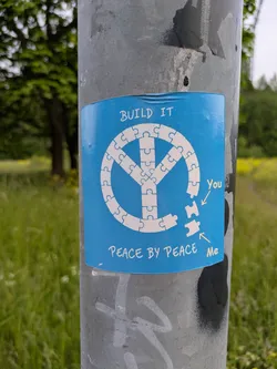 Street sticker A blue sticker affixed to a metal pole features a peace symbol constructed from jigsaw puzzle pieces. Two pieces are separate from the symbol, labeled "You" and "Me", encouraging individual action towards building peace. The text "BUILD IT" appears above the symbol, and "PEACE BY PEACE" below it.