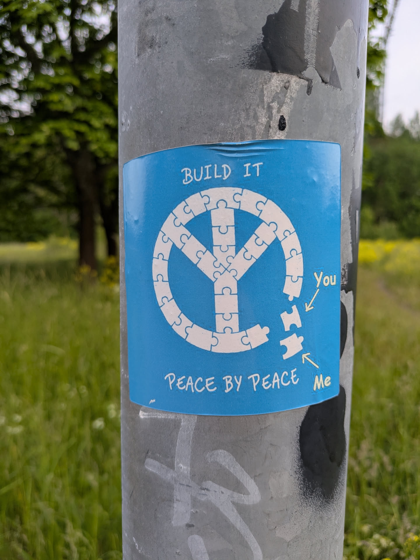 A blue sticker affixed to a metal pole features a peace symbol constructed from jigsaw puzzle pieces. Two pieces are separate from the symbol, labeled "You" and "Me", encouraging individual action towards building peace. The text "BUILD IT" appears above the symbol, and "PEACE BY PEACE" below it.