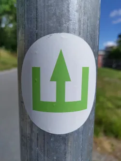 Street sticker A white circular sticker affixed to a gray metal pole. The sticker features a bright green symbol; an upward-pointing arrow within a rectangular frame. The design is simple and bold. The sticker appears to be recently placed and in good condition.