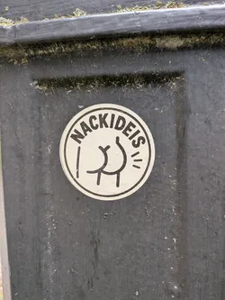 Street sticker A round white sticker with a black border. It displays the arched German text 'NACKIDEIS' above a simple black line drawing of a naked rear end.