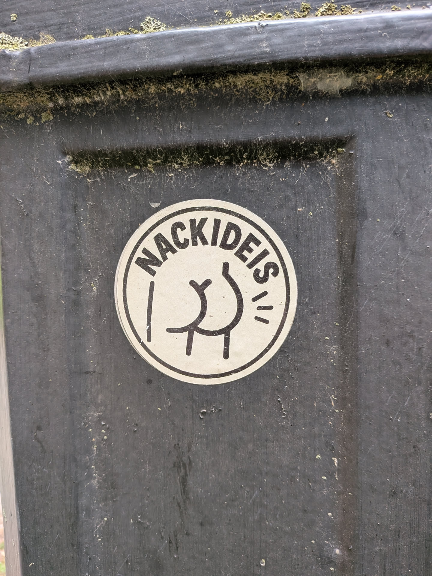 A round white sticker with a black border. It displays the arched German text 'NACKIDEIS' above a simple black line drawing of a naked rear end.