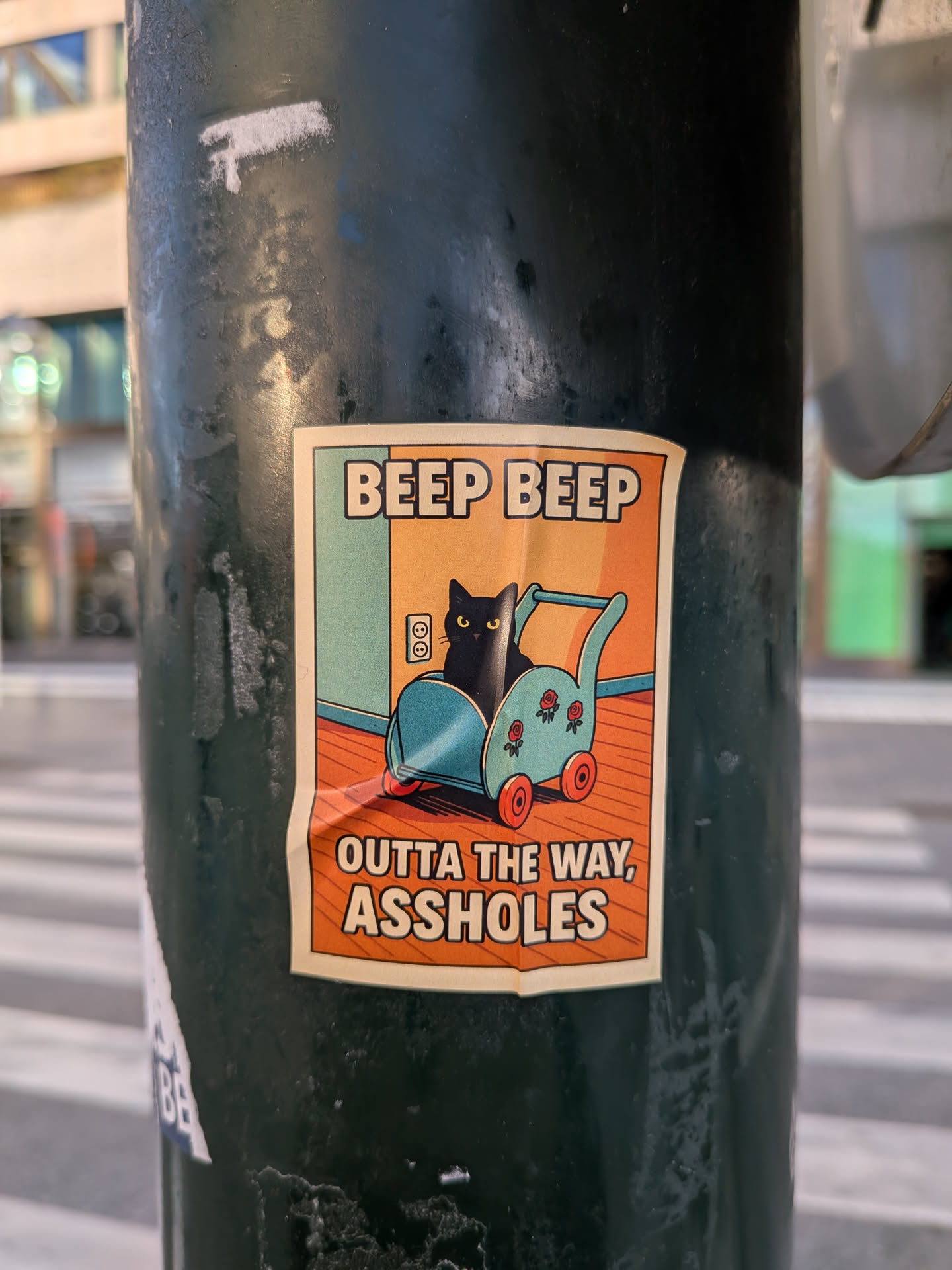 The sticker is rectangular with rounded corners and a cream-colored border. The central image depicts a black cat with yellow eyes peeking out from a teal-colored baby carriage. The carriage has red wheels and is decorated with two small red rose emblems. The background is split into two sections: the upper part is a light orange-yellow, and the lower part is a light blue. To the left of the cat, there's a depiction of a wall with an electrical outlet. The bottom of the sticker features bold, orange text on a cream background. The overall style is reminiscent of vintage illustrations.