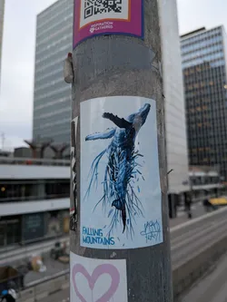 Street sticker The sticker features a striking illustration of a whale, predominantly rendered in various shades of deep and light blue. The whale is depicted in a dynamic, almost falling or diving posture, with its fins and flukes spread. Below and around the whale, numerous abstract, elongated, and somewhat jagged blue elements resembling roots, tendrils, or fragmented ice/mountains create a sense of motion and disintegration. The background of the sticker is a light, almost white-blue, allowing the darker blue elements to stand out prominently. The overall style is illustrative and surreal, with a distinct artistic signature in the bottom right corner.