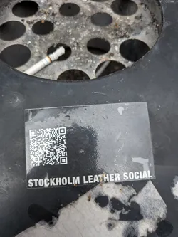 Street sticker A rectangular sticker, predominantly black, featuring a QR code on the left side and white text on the right. The QR code is clearly defined with black and white squares. Below the QR code, the text 'STOCKHOLM LEATHER SOCIAL' is printed in bold, sans-serif white capital letters. The sticker appears to be slightly glossy and has some minor scuffs and what looks like a small water droplet or smudge on its surface.