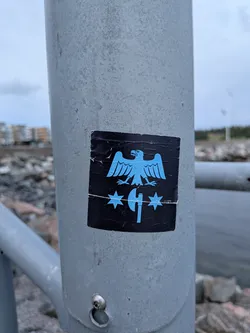 Square black sticker on a metal pole. Features a light blue graphic of a stylized eagle with spread wings above a vertical axe, flanked by two six-pointed stars. A horizontal crease is visible across the lower section.