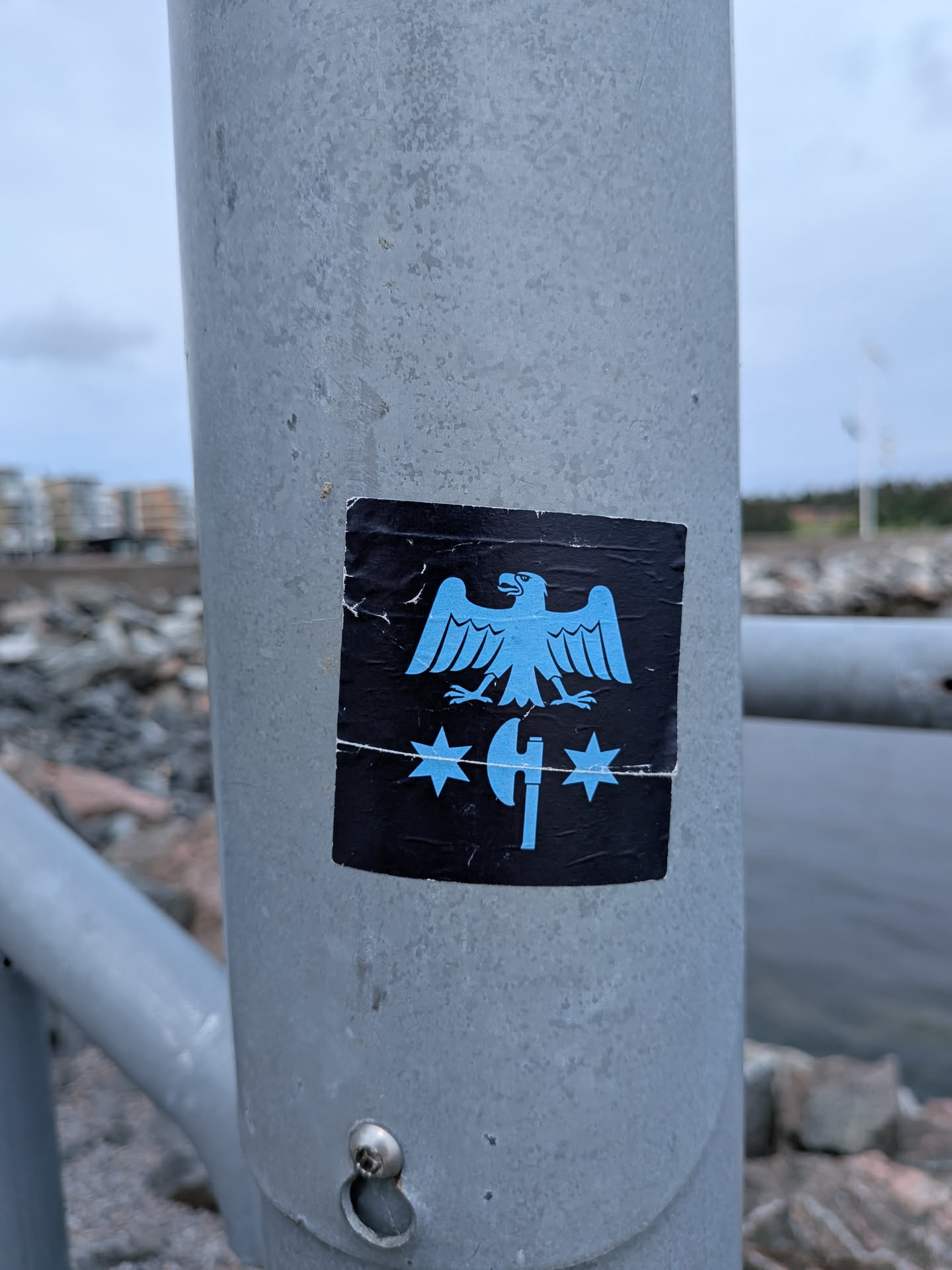 Square black sticker on a metal pole. Features a light blue graphic of a stylized eagle with spread wings above a vertical axe, flanked by two six-pointed stars. A horizontal crease is visible across the lower section.
