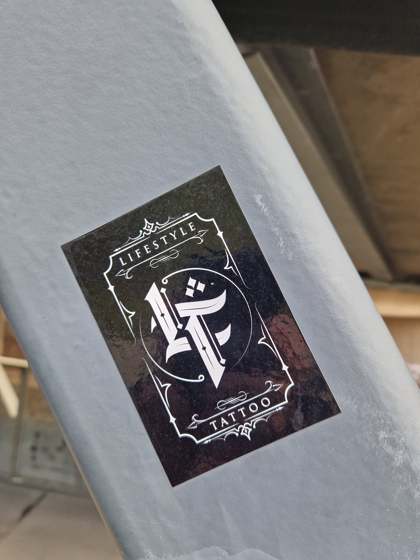A rectangular sticker with a glossy black background. It features a white ornate border with decorative flourishes at the top and bottom. Inside the border, the word 'LIFESTYLE' is written at the top and 'TATTOO' at the bottom. The center contains a circular frame enclosing a stylized, gothic-style monogram of the letters 'L' and 'T'.