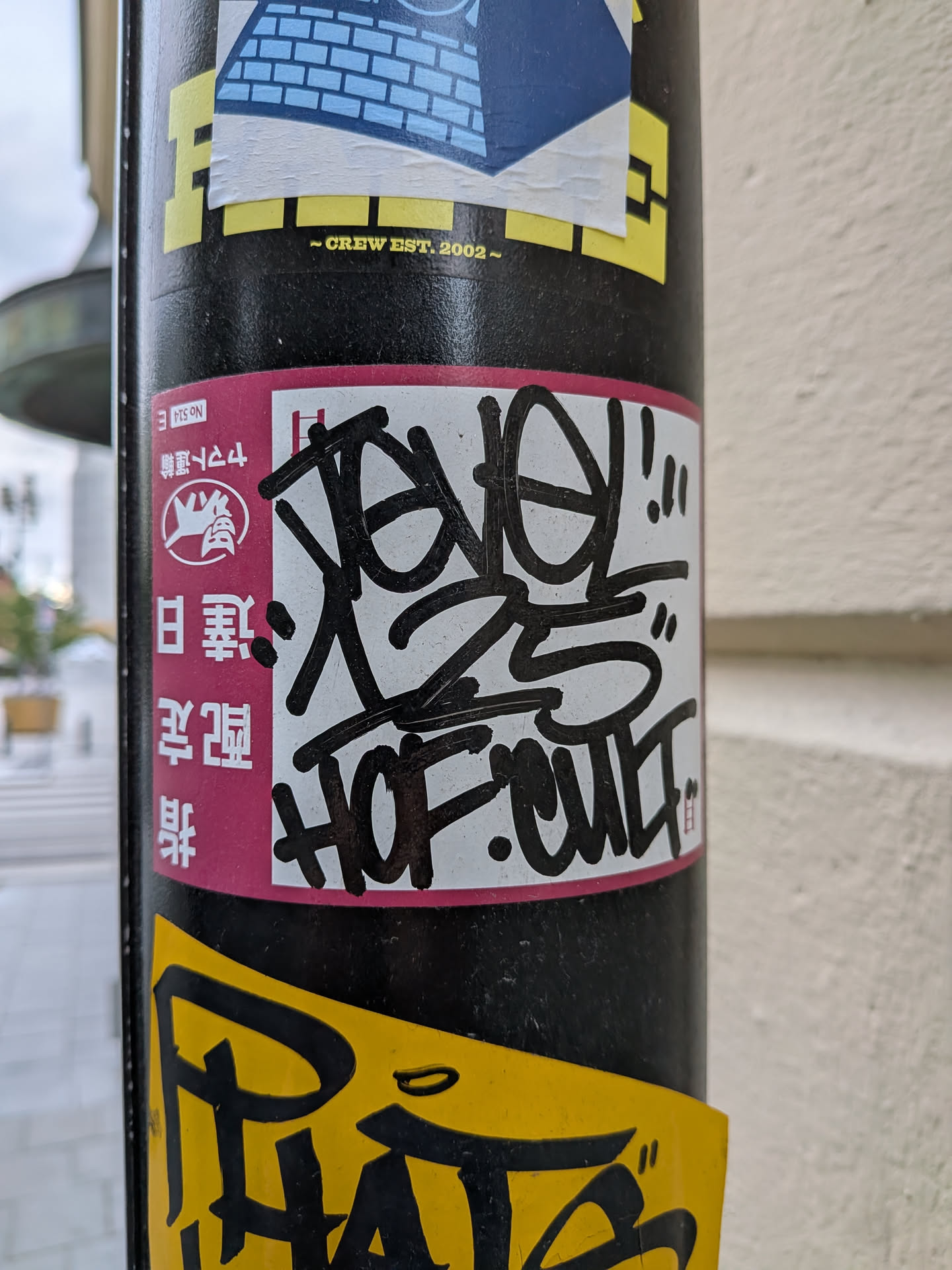 A rectangular sticker with a pink left margin containing Japanese text and a black cat logo. The white center features a bold black marker graffiti tag reading 'REVEL 25' and 'HOF CULT'.
