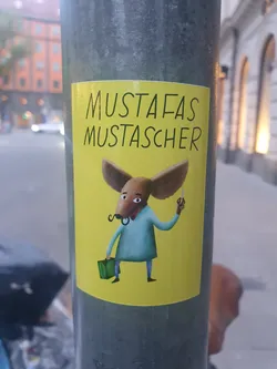 Street sticker A yellow sticker featuring an illustration of an anthropomorphic animal, possibly a mouse or a similar creature, holding scissors and a briefcase. The animal has a mustache and is dressed in a light teal shirt. The text "MUSTAFAS MUSTASCHER" is written at the top of the sticker.