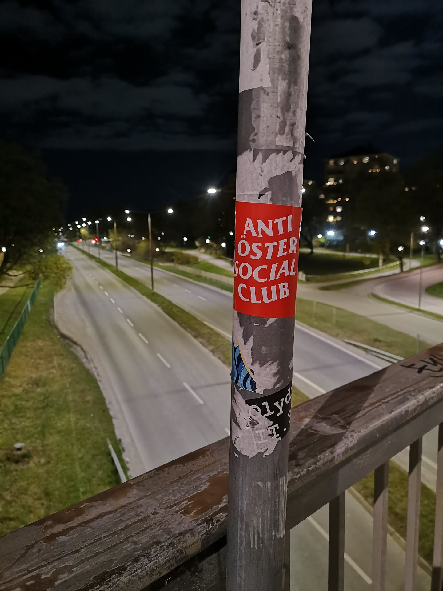 A rectangular sticker with a vibrant red background and clear white sans-serif text. The text is stacked, reading 'ANTI' at the top, 'ÖSTER' in the middle, and 'SOCIAL CLUB' at the bottom, all in uppercase letters. The sticker is affixed to a weathered grey metal pole, showing minor signs of wear and tear around its edges, consistent with outdoor placement.