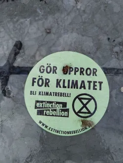 Street sticker A round light green sticker with black text and the Extinction Rebellion logo. The text translates to "Rebel for the climate! Become a climate rebel!"