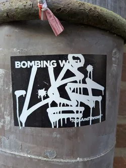 The sticker is a rectangular, horizontally oriented design with a matte black background. Prominently featured in the center is a complex, stylized graffiti-style lettering in white, characterized by thick strokes and numerous paint drips extending downwards, giving the impression of fresh, wet paint. The white lettering appears to form an abstract tag or throw-up. In the upper left corner, in a clear, sans-serif white font, are the words 'BOMBING W'. In the bottom right corner, also in white, is the text 'TagsAndThrows™'.