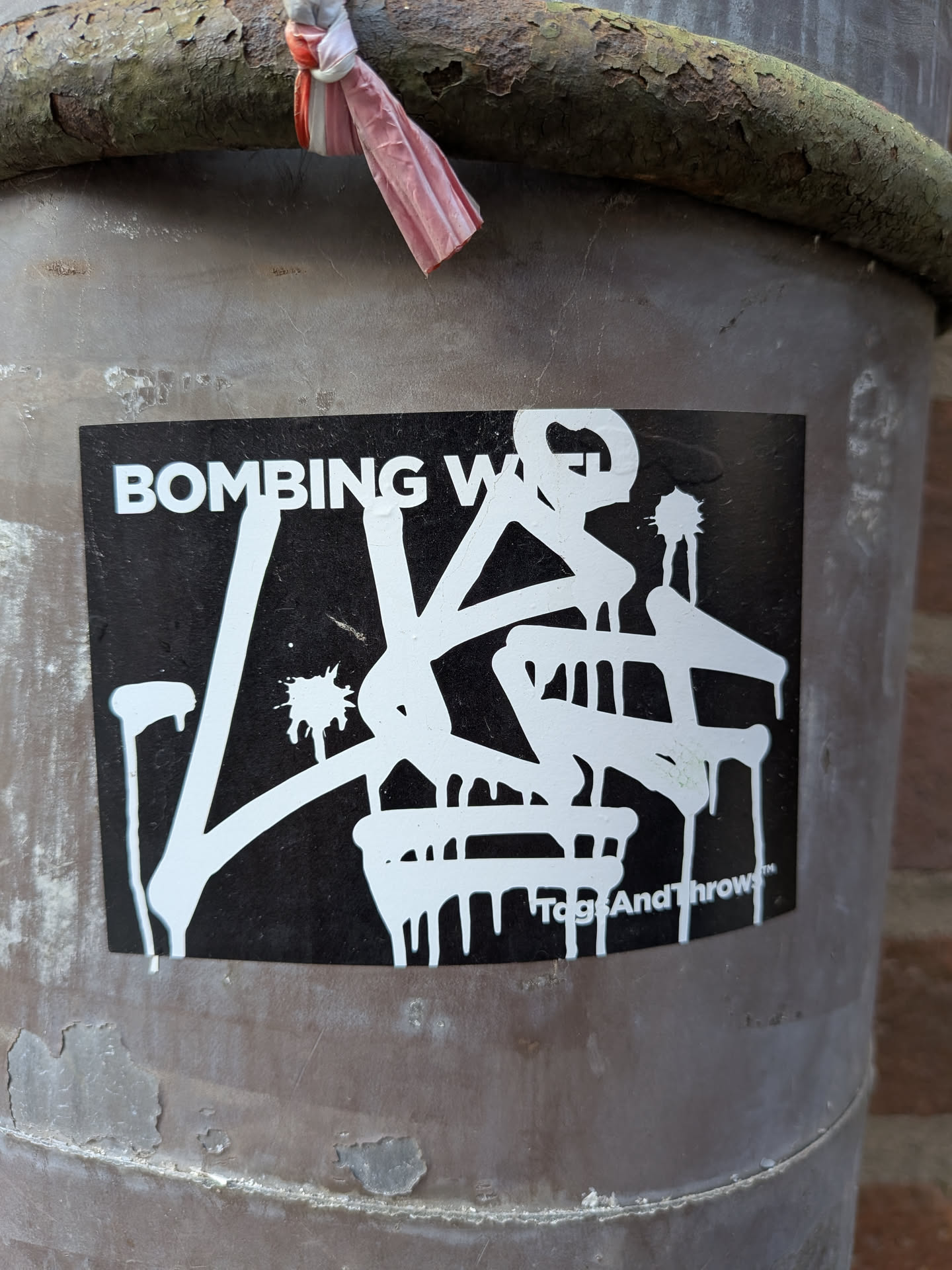 The sticker is a rectangular, horizontally oriented design with a matte black background. Prominently featured in the center is a complex, stylized graffiti-style lettering in white, characterized by thick strokes and numerous paint drips extending downwards, giving the impression of fresh, wet paint. The white lettering appears to form an abstract tag or throw-up. In the upper left corner, in a clear, sans-serif white font, are the words 'BOMBING W'. In the bottom right corner, also in white, is the text 'TagsAndThrows™'.