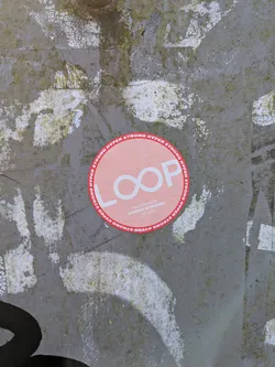 Street sticker A circular sticker featuring a light peach-orange background with a prominent red border. The border contains white, uppercase text that repeatedly reads 'HYPER STRONG'. In the center, the word 'LOOP' is displayed in large, bold white letters, with the two 'O's stylized to form an infinity symbol. Below 'LOOP', smaller white text reads 'HOT PEACH', followed by 'HYPER STRONG', and then '18+' and 'HOT!' in even smaller text, with '18+' enclosed in a small circle. The sticker appears to be in good condition, contrasting with the heavily weathered and dirty grey surface it's affixed to.