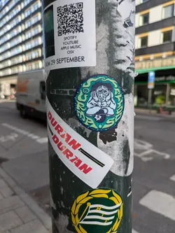 Street sticker A round sticker featuring a drawing of a seated figure with a thoughtful expression, surrounded by vibrant colors and swirling patterns. The style is reminiscent of 70s and 80s psychedelic art. The sticker is applied to a pole along with other stickers, including a Duran Duran band sticker and a QR code linking to various music platforms.