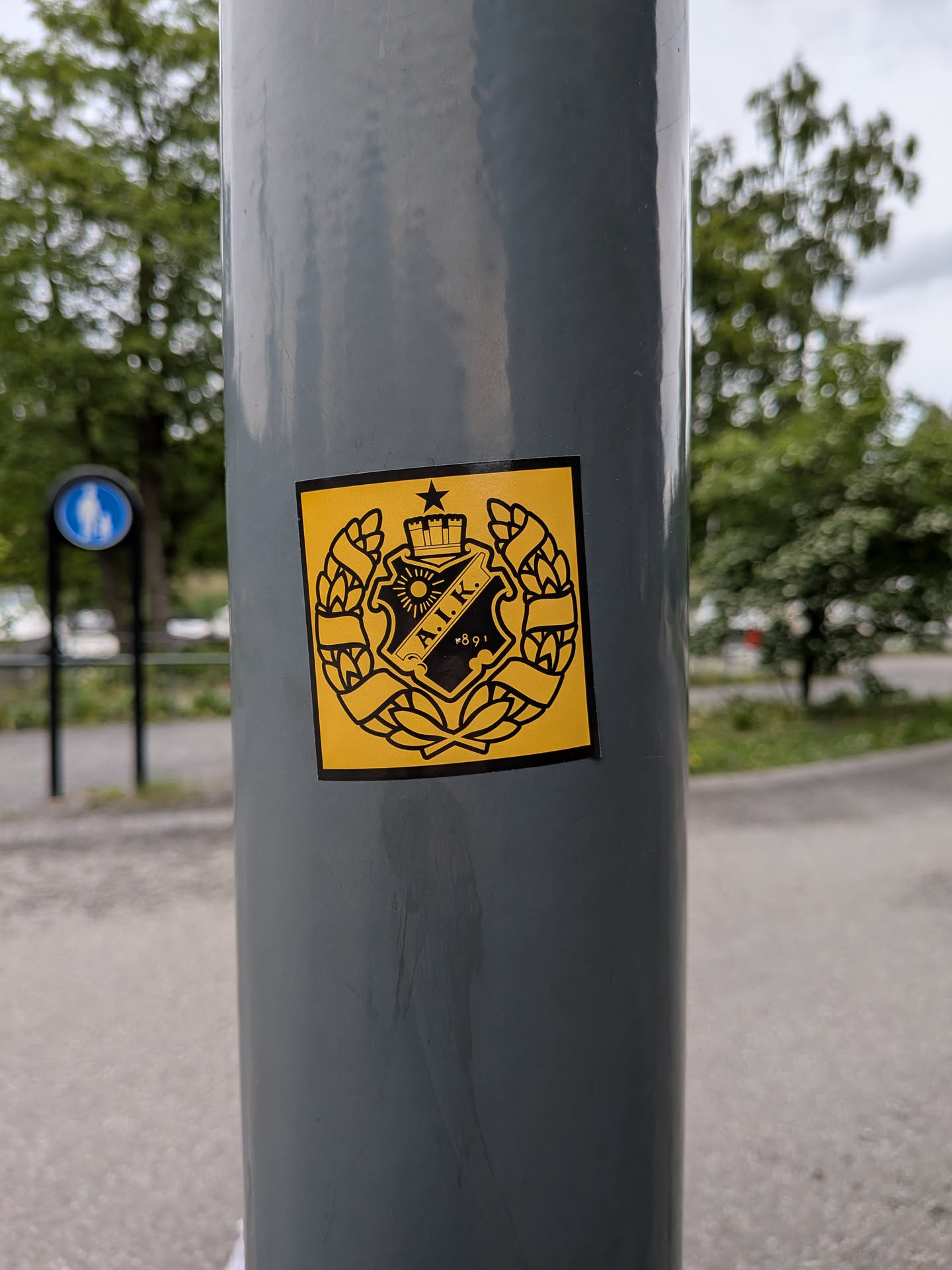 A square yellow sticker featuring the black crest of the Swedish sports club AIK. The crest includes a central shield with 'A.I.K.' and '1891', topped by a crown and star, and enclosed in a wreath.
