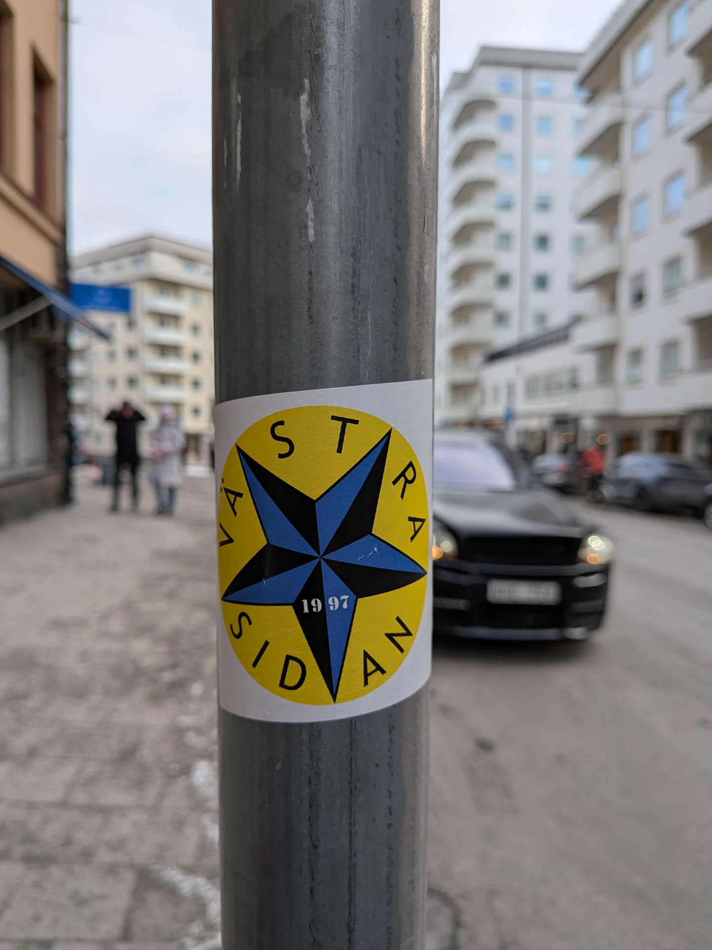 Street sticker A circular sticker with a bright yellow background and a white outer border, affixed to a grey metal pole. The central design features a prominent five-pointed star, rendered in blue with black outlines and internal divisions, giving it a multi-faceted, almost compass-like appearance. Around the star, the words "VÄSTRA SIDAN" are written in black capital letters, curving along the yellow background. "VÄSTRA" is on the left side, and "SIDAN" is on the right. At the bottom point of the blue star, the year "1997" is inscribed in black numerals. The sticker shows minor signs of wear, with slight lifting at some edges.