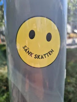 Street sticker A yellow smiley face sticker is affixed to a gray pole. The sticker has the text "Sänk Skatten", which translates to "Lower the Taxes" in English, written in a curved arc below the smiley face's smile. The sticker is simple, with bold black outlines for the eyes and smile.