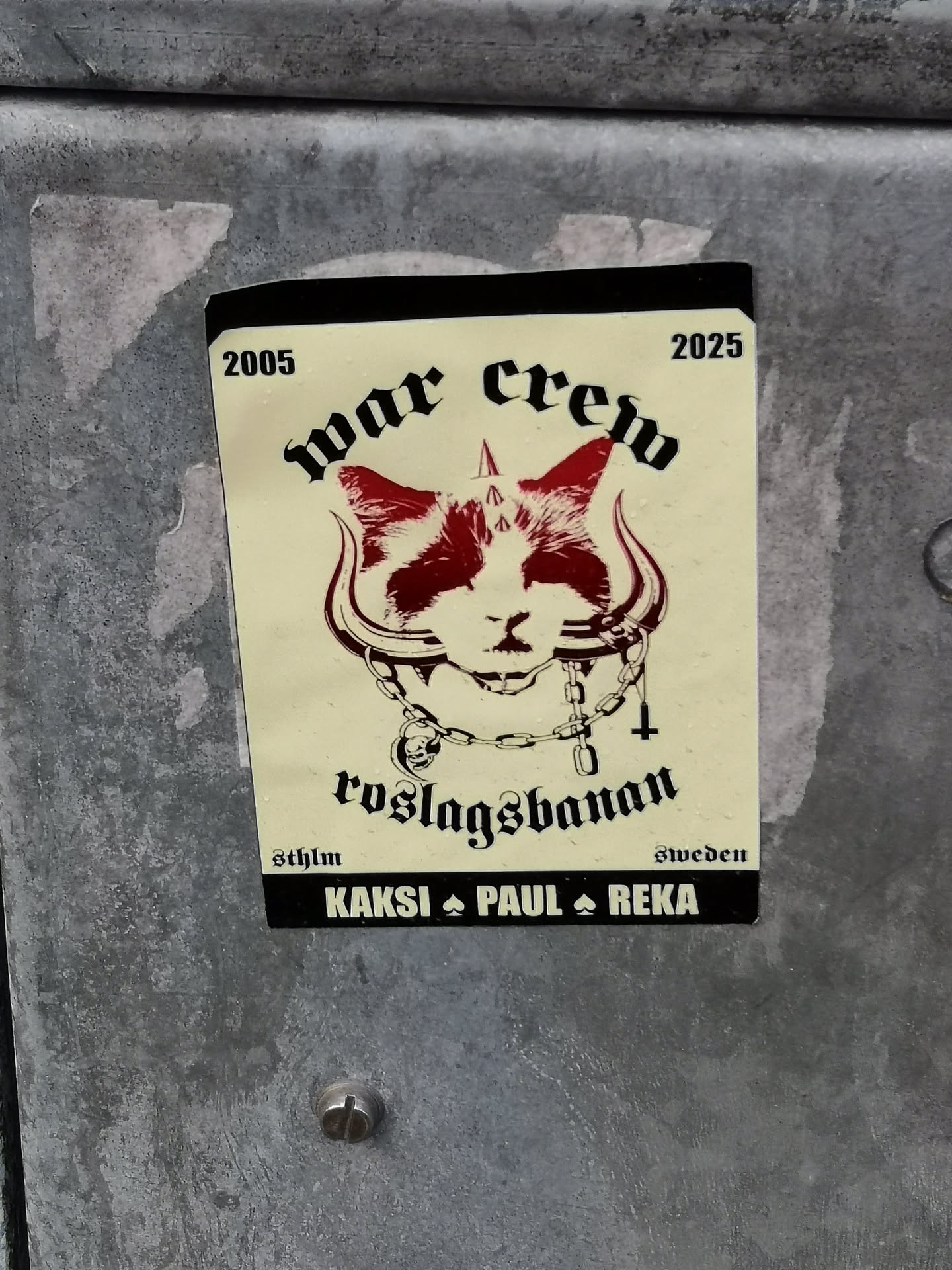 The sticker features a stylized, somewhat aggressive cat's head with prominent, curved horns extending from its temples. The cat's face is predominantly white with dark red, almost blood-like, markings around its eyes and ears. It wears a thick, dark chain collar adorned with a small skull charm and an inverted cross pendant. Above the cat, the words 'war crew' are written in a bold, gothic-style font, flanked by the years '2005' on the left and '2025' on the right. Below the cat, 'roslagsbanan' is inscribed in a similar gothic script. The bottom section of the sticker includes 'sthlm' on the left and 'sweden' on the right, with a black bar at the very bottom containing the names 'KAKSI ♠ PAUL ♠ REKA' separated by spade symbols. The background is an off-white or cream color, and the overall design has a dark, punk, or metal subculture aesthetic.