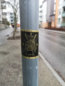 Street sticker A rectangular sticker with a black background and gold-colored design, wrapped around a wet, grey metal pole. The central motif is a circular emblem featuring a detailed gold outline of a penny-farthing bicycle. Behind the bicycle, two swords are crossed. The emblem is framed by a thin gold border at the top and bottom, adorned with small black dots resembling rivets. Partial gold text is visible around the central design: 'A' on the left, 'K' on the right, and '1891' clearly at the bottom. The sticker shows signs of wear and moisture, with visible wrinkles and water droplets.