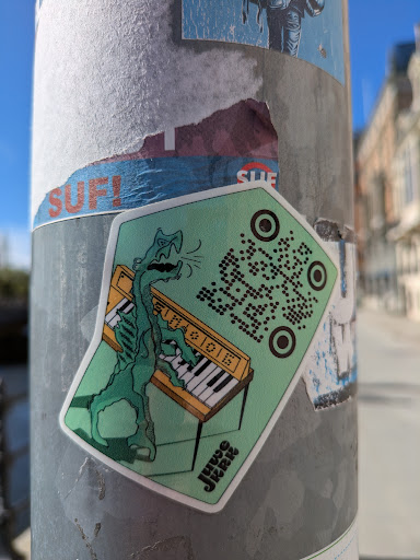 A light-green, pentagon-shaped sticker on a pole features a stylized illustration of a tall, slender, green monster with a scaly texture playing a tan-colored piano. The piano has black and white keys. The sticker includes a QR code on the top right. The name 'JUWO JRRR' is printed in stylized font at the bottom of the sticker.