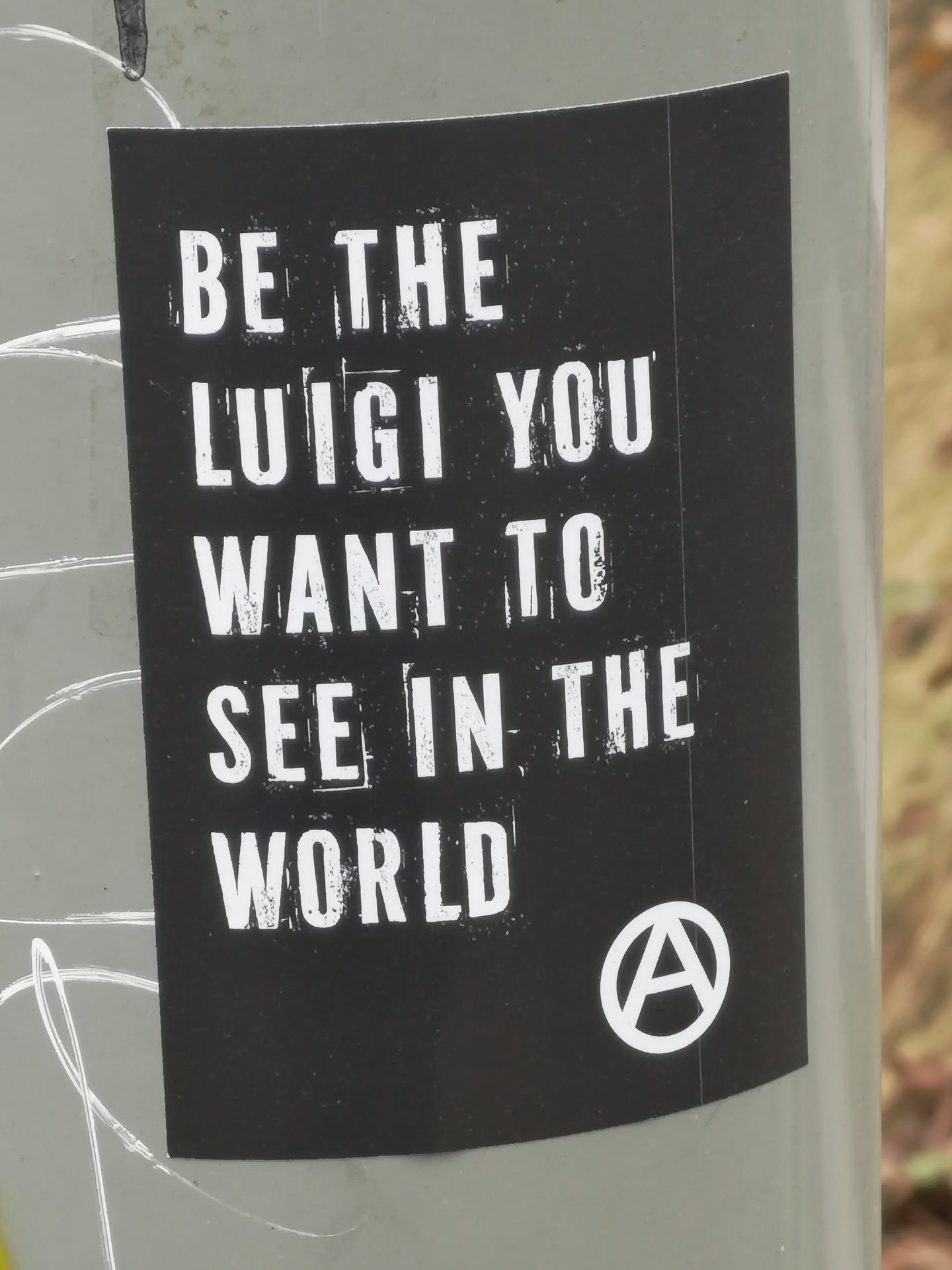 A rectangular black sticker with white, distressed, uppercase text that reads 'BE THE LUIGI YOU WANT TO SEE IN THE WORLD'. In the bottom right corner, there is a white circle containing a stylized 'A' symbol, commonly recognized as the anarchy symbol. The sticker has a slightly worn appearance, with some white specks visible on the black background, suggesting texture or age. It is affixed to a grey pole, with some white graffiti marks visible around its edges.