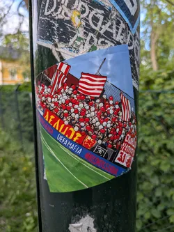 Street sticker The sticker illustrates a packed football stadium from a low angle, showing a large crowd of fans in the stands. The fans are depicted in a cartoonish style, many holding red and white balloons or wearing red and white attire. Several large flags with red and white horizontal stripes are prominent among the crowd. The sky above is light blue, and a green football pitch with white lines is visible in the foreground. A blue banner runs along the bottom edge of the crowd, featuring white text. Another yellow banner with red text is visible above it. On the right, a white banner with red text is partially visible, and below the main crowd, a small black banner with a skull and crossbones is present.