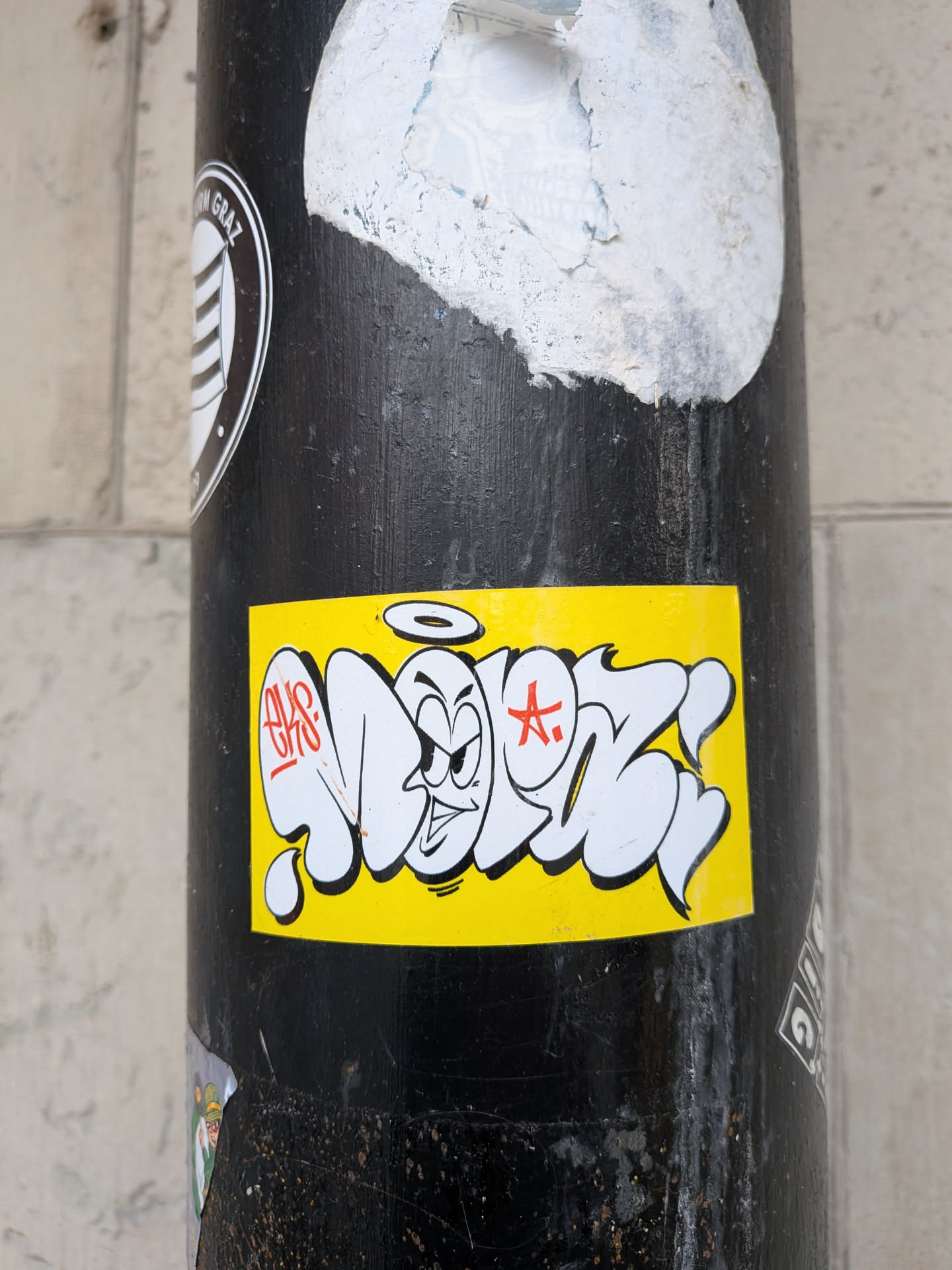The sticker is rectangular with a bright, solid yellow background. The central design consists of white, thick, bubble-style graffiti letters outlined in black. Integrated into these letters is a cartoon character's face with a mischievous or angry expression, featuring furrowed brows, wide eyes, and a downturned mouth. A simple black halo floats above the character's head. To the left of the character, the letters 'EKS' are written in red, stylized script. To the right, a red, stylized 'A.' with a star-like element is visible. The overall aesthetic combines graffiti throw-up style with cartoon illustration.