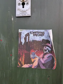 Street sticker A square-shaped sticker featuring a stylized illustration of a skeleton seated, wearing a wide-brimmed purple hat and matching attire. The skeleton holds a silver mate gourd with a bombilla (metal straw) in its skeletal hands. Faint text 'I AM A VILLAIN TO MY B...' is visible on the gourd. The background depicts a desolate urban landscape with ancient-looking stone ruins on the left, brick walls with a reddish glow suggesting fire or sunset, and a modern building in the distance under a pale, textured sky. The prominent title 'SOMETHING DEAD' is displayed at the top in a distressed, blocky font, with 'SOMETHING' in a lighter blue and 'DEAD' in a darker blue/black. The overall color scheme is muted, dominated by purples, blues, reds, and browns, creating a somber yet intriguing atmosphere. The sticker shows signs of wear, with a slightly peeled corner on the top right.