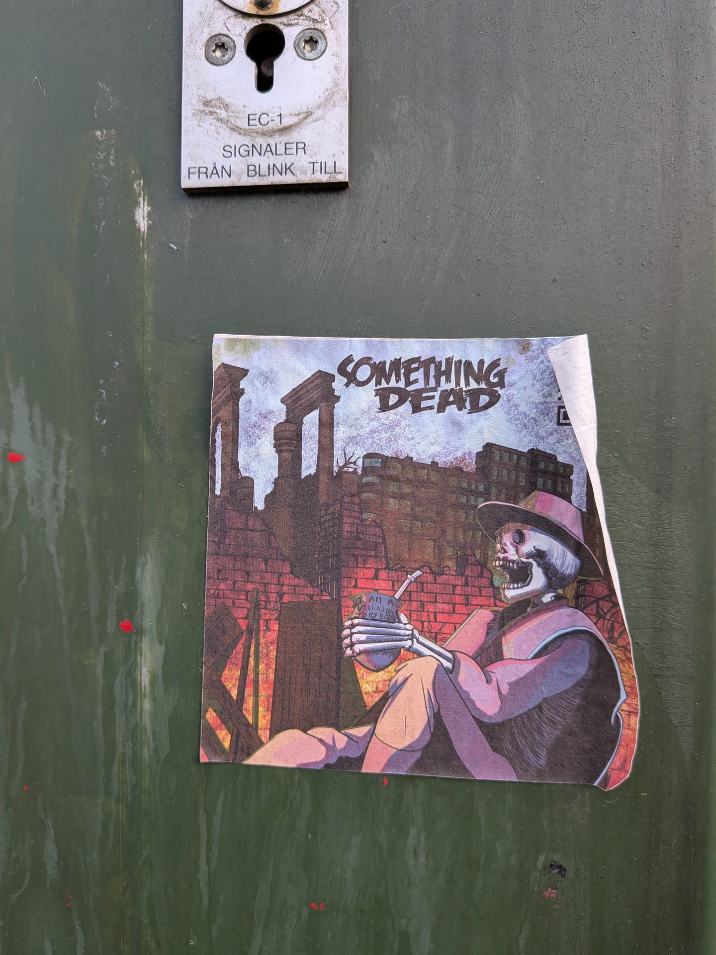 A square-shaped sticker featuring a stylized illustration of a skeleton seated, wearing a wide-brimmed purple hat and matching attire. The skeleton holds a silver mate gourd with a bombilla (metal straw) in its skeletal hands. Faint text 'I AM A VILLAIN TO MY B...' is visible on the gourd. The background depicts a desolate urban landscape with ancient-looking stone ruins on the left, brick walls with a reddish glow suggesting fire or sunset, and a modern building in the distance under a pale, textured sky. The prominent title 'SOMETHING DEAD' is displayed at the top in a distressed, blocky font, with 'SOMETHING' in a lighter blue and 'DEAD' in a darker blue/black. The overall color scheme is muted, dominated by purples, blues, reds, and browns, creating a somber yet intriguing atmosphere. The sticker shows signs of wear, with a slightly peeled corner on the top right.