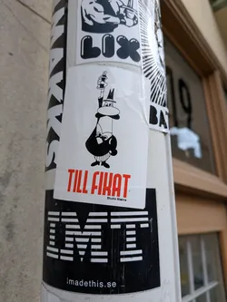 Street sticker The sticker features a whimsical, black and white cartoon character on a white rectangular background. The character, depicted in a minimalist style, wears a top hat and a suit, has a prominent nose, and a small beard. It holds a spray paint can upright like a scepter or staff. Below the character, in large red capital letters, is the text 'TILL FIKAT', with 'Studio Stains' written in smaller black text underneath. The overall aesthetic is clean and graphic.