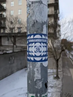 Street sticker The sticker is rectangular, wrapped around a metal pole, and features a striking design in dark blue and white. At the top, the word "KOMMANDOBRYGGAN" is written in white capital letters against a blue background. The main body of the sticker showcases a repeating geometric pattern composed of white semi-circles and quarter-circles on a blue background, creating a tessellated, almost wave-like or mosaic effect. Centered within this pattern is a white square containing a blue crest or emblem. The crest features a stylized animal (possibly a lion or griffin) and the letters "I.F.K." above it. The overall appearance is clean, graphic, and has a strong visual identity.