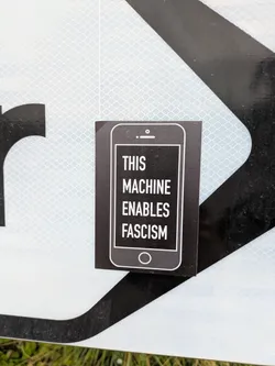 Street sticker A rectangular black sticker featuring a minimalist white outline of a smartphone. Inside the screen area of the smartphone, the text 'THIS MACHINE ENABLES FASCISM' is displayed in white, sans-serif capital letters, stacked vertically. The sticker is adhered to a reflective white and black surface, likely a road sign.
