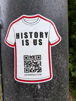 Street sticker A white, t-shirt-shaped sticker outlined in red and black. It displays the bold text 'HISTORY IS US', a QR code, and the website 'AEWHOLE.COM' on a dark textured pole.