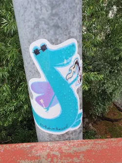 A sticker featuring a teal crocodile with sunglasses, fairy wings, and a wand. The artist's signature is visible at the bottom: "Filio Rahim Hansson"