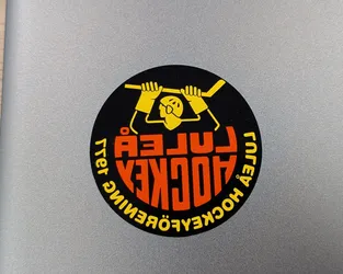 Street sticker A circular sticker featuring a bold, graphic design in black, orange, and yellow. The center depicts a stylized, cartoonish figure in yellow, possibly a hockey player or goalie, holding a hockey stick horizontally above its head. The figure has simple facial features and large gloves. The word 'HOCKEY' is prominently displayed in large orange letters across the center, with 'LULEÅ' in smaller orange letters directly above it. Around the outer edge, in yellow, are the words 'ÅTER' (top left) and 'HOCKEYÖVNING' (bottom right), both of which are mirrored as a stylistic choice, adding to the unique retro aesthetic.