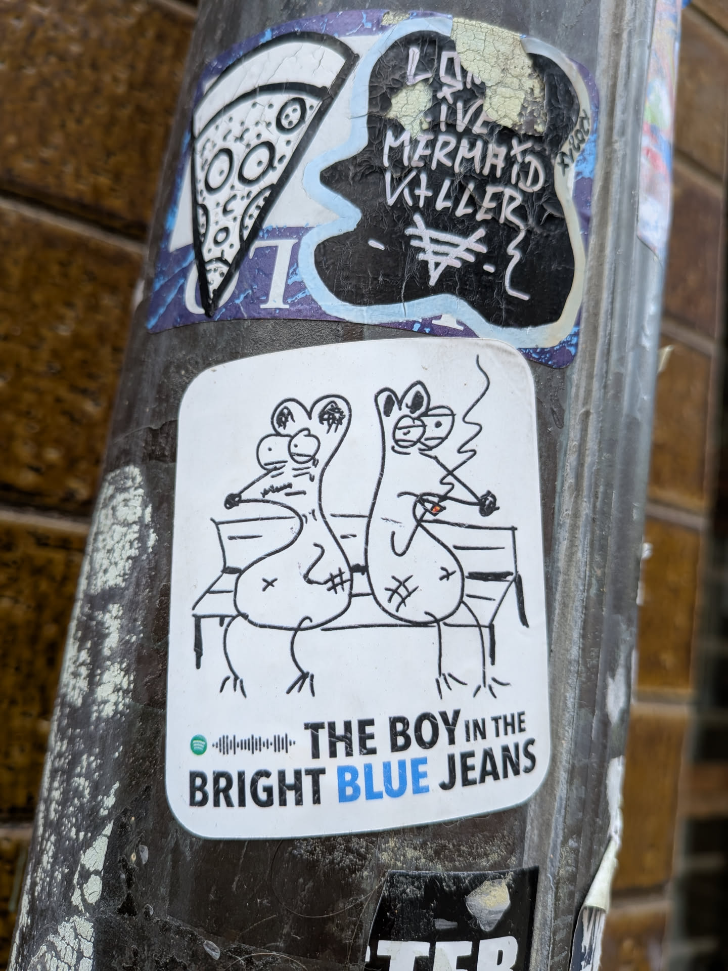 The central sticker is a white, rectangular shape with rounded corners, featuring a black line-art drawing on a white background. It depicts two stylized, somewhat mouse-like or rat-like cartoon figures with large ears and prominent snouts, sitting side-by-side on a park bench. The figure on the right is smoking a cigarette, indicated by a small red tip and a wavy line representing smoke. Both figures have simple, somewhat dazed or relaxed expressions. Below the drawing, there's a small green Spotify logo followed by a black sound wave graphic. Underneath this, the text "THE BOY IN THE BRIGHT BLUE JEANS" is written in black, with the word "BLUE" highlighted in a vibrant blue color. The overall style is whimsical and minimalist.