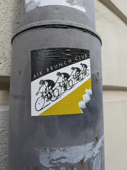 Street sticker A square sticker featuring a dynamic graphic of four cyclists in a line, depicted in black silhouettes against a white and yellow background. The top portion of the sticker is black, with the text 'AIK BRUNCH CLUB' written in white, uppercase letters diagonally across it. Below this, a diagonal line separates a white section from a yellow section, creating a sense of movement. The cyclists are rendered in a minimalist, illustrative style, suggesting a team or club. The sticker shows signs of wear, with wrinkles and some peeling, indicating it's a physical sticker adhered to a surface.