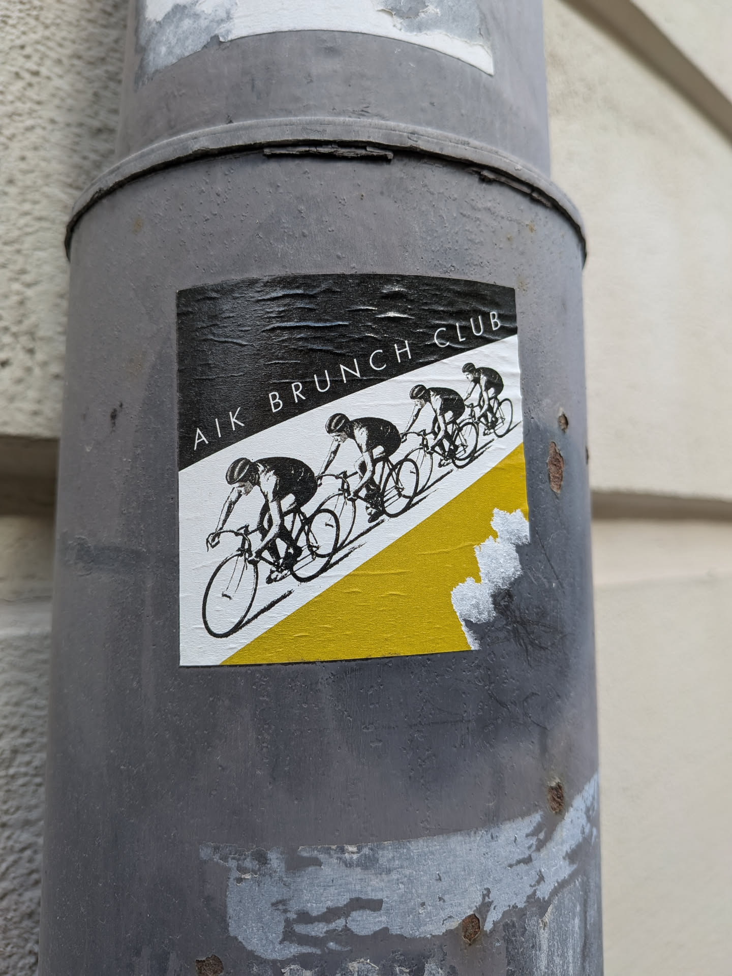 A square sticker featuring a dynamic graphic of four cyclists in a line, depicted in black silhouettes against a white and yellow background. The top portion of the sticker is black, with the text 'AIK BRUNCH CLUB' written in white, uppercase letters diagonally across it. Below this, a diagonal line separates a white section from a yellow section, creating a sense of movement. The cyclists are rendered in a minimalist, illustrative style, suggesting a team or club. The sticker shows signs of wear, with wrinkles and some peeling, indicating it's a physical sticker adhered to a surface.