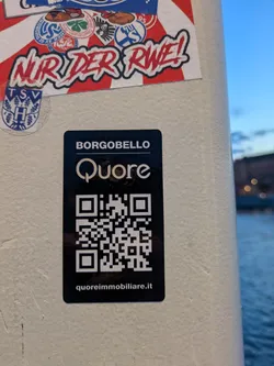 Street sticker The sticker is predominantly black with white and light gray text and graphics. At the top, in white capital letters, it reads 'BORGOBELLO'. Below this, a stylized white 'Q' with a circular loop forms the logo for 'Quore'. The majority of the sticker is occupied by a scannable QR code in black and white. At the bottom, in smaller white text, is the website 'quoreimmobiliare.it'. The sticker has a glossy finish.