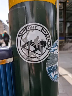 Street sticker The sticker is circular with a white border. Inside, it features a black silhouette of a person riding a horse. The horse appears to be in motion, with its legs extended. Above the figures, the text "SUPPORT" is written in uppercase black letters, arching along the top of the circle. To the right, also in an arch, is the text "HRAVN MC". The design is primarily black and white, with some distressed silver-gray shading and cracks visible on the sticker's surface, indicating wear and tear. A smaller, partially obscured sticker with blue and white elements is visible to the right of the main sticker.