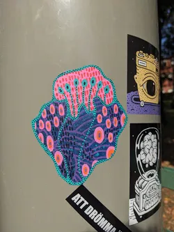 Close-up view of a vibrant sticker featuring a stylized coral or sea anemone with pink, purple, teal, and orange patterns. The artwork is detailed and intricate, with a psychedelic feel.  It's adhered to a grey surface alongside other stickers.