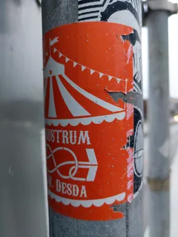 An orange sticker featuring a simple illustration of a circus tent with the word 'lustrum' and a symbol below it.  The sticker shows some wear and tear, suggesting it has been outdoors for a while. The bottom features the text 'N. DESDA'.