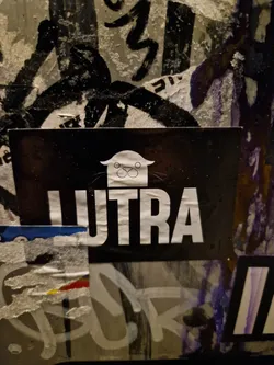 Street sticker A black and white sticker with the word "UTRA" in bold white lettering.  Above the word is a small, simple illustration of a creature that resembles a stylized cat or otter with a house-like shape on its head. The sticker is affixed to a wall covered in other graffiti and stickers.
