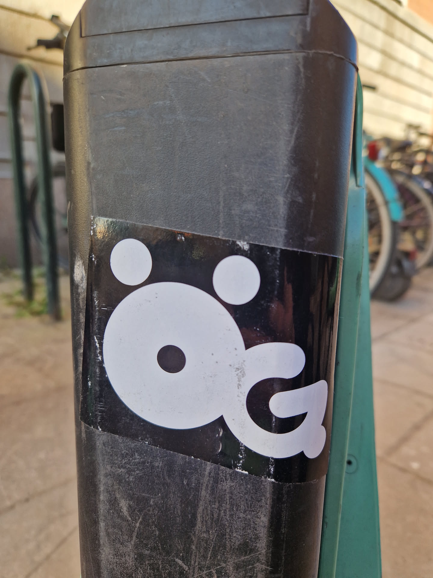 The sticker features a minimalist, abstract character design in white against a rectangular black background. The character consists of a large central white oval shape, which could represent a head or body, with a small black circle in its center. Above this central shape are two smaller white circles, positioned like eyes. To the right, a distinctive white shape resembling a stylized letter 'G' or a curled limb extends from the main body. The sticker shows signs of wear and slight peeling at the edges, indicating it's a physical sticker applied to a textured, dark surface.