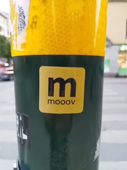 A gold square sticker with a black 'm' and the word 'mooov' underneath.  It is affixed to a dark green pole. The sticker has a clean, minimalist design.